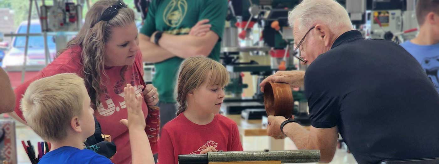 a young girl and her family watch a demonstration on how to make a wooden bowl on a lathe